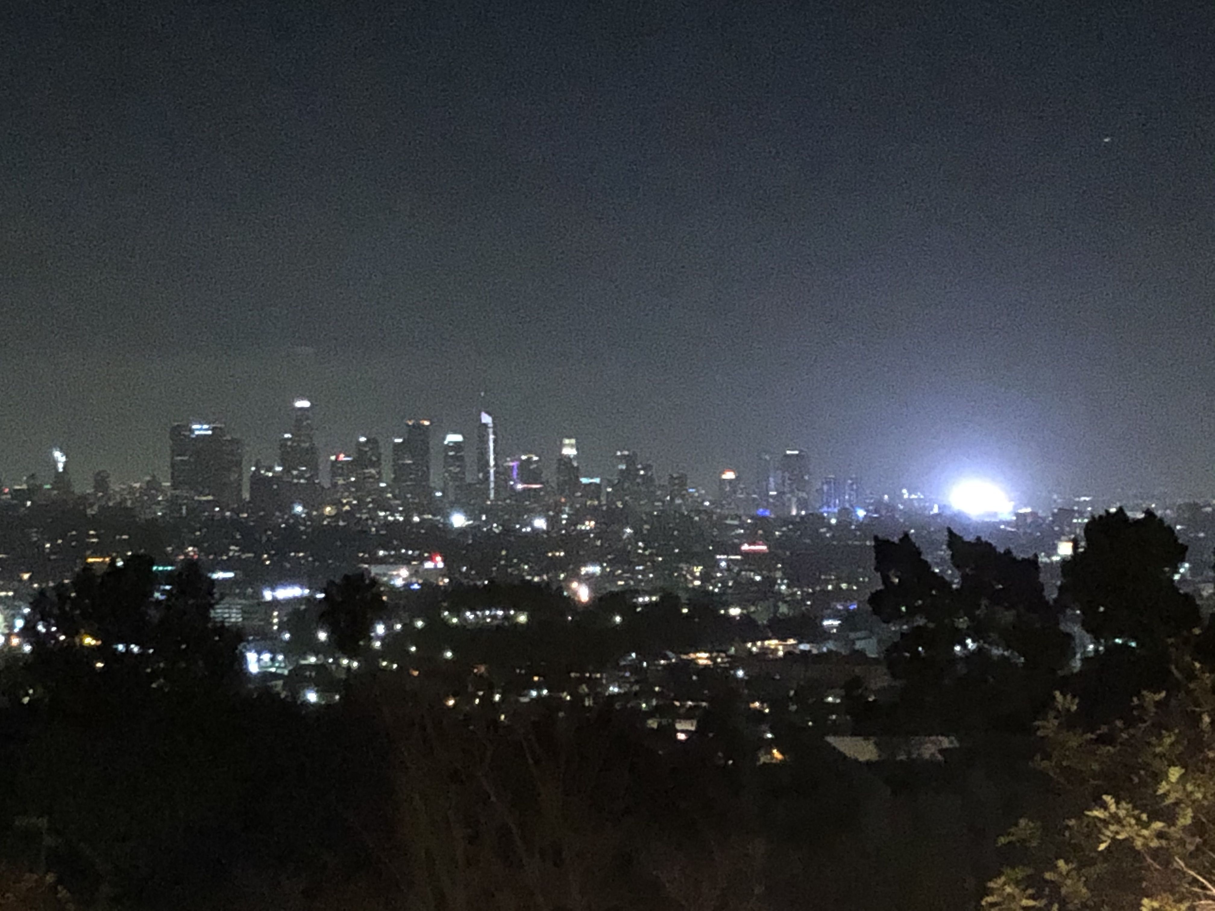 A photo taken at night over Los Angeles with many illuminated buildings. In the distance, an extraordinarily bright light overpowers the scene. It is an advertising billboard.