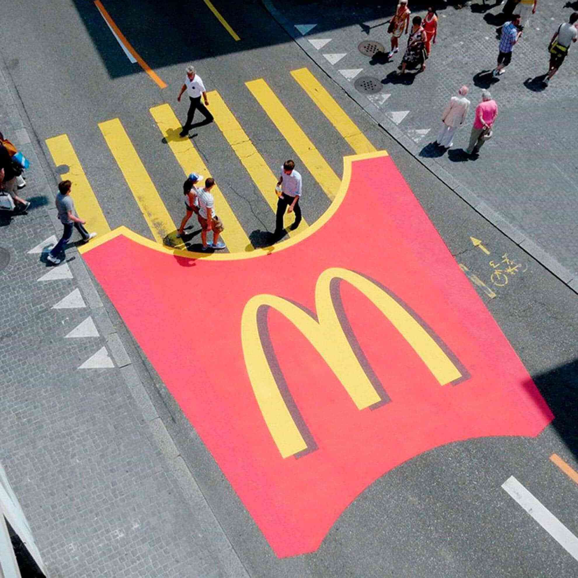 A photo taken from a very high angle looks down at the street, showing that a pedestrian crosswalk has been turned into a mcdonalds advertisement. The ordinary yellow crosswalk lines now represent french fries, coming out of a large red mcdonalds fry cup.