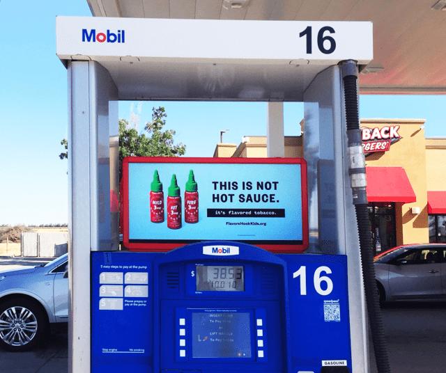 A photo of a gas pump at a gas station. On top of the pump is a screen showing an ad for hot sauce.