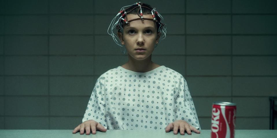 Screenshot from Stranger Things. Eleven, wearing a medical gown and crown of brain activity monitors, sits at a desk with a can of Coke.