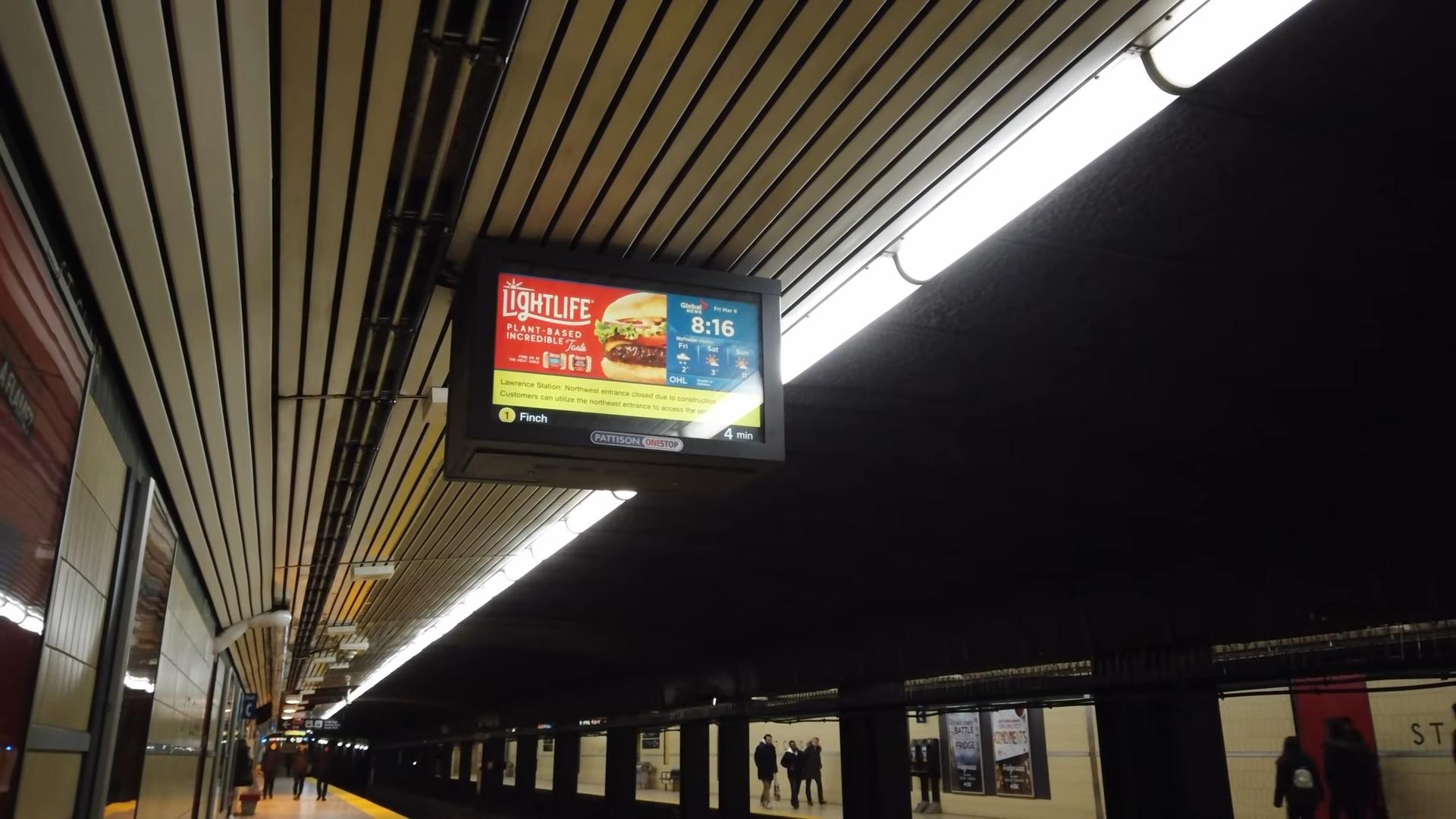 A photo taken inside a subway station. A digital screen hangs from the ceiling. The left two thirds of the screen are an advertisement for a restaurant. The right one third of the screen shows the time of the next arriving train and the weather.