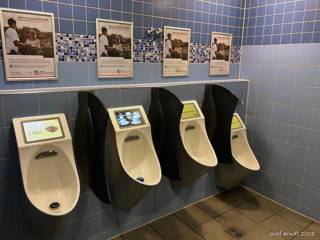 A photo of four urinals in a public restroom. Each of the urinals has a digital screen integrated into the body of the urinal, angled upward towards the guest. The screens show products and branding.